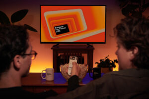A silhouette of two adults on a couch. The person on the left points a remote control at a TV screen with Festival branding.